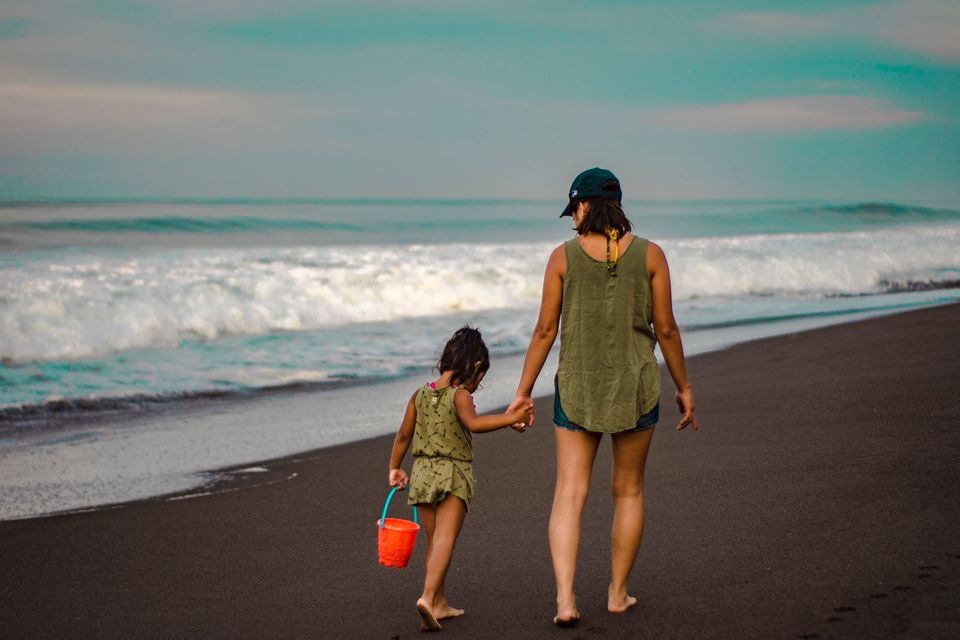 A mother and her daughter walking hand in hand along a beach at dusk, seen from behind